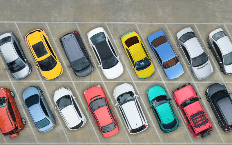 Aerial view of a parking lot with cars parked in two rows, featuring various colors like yellow, green, red, and blue, on a concrete surface.