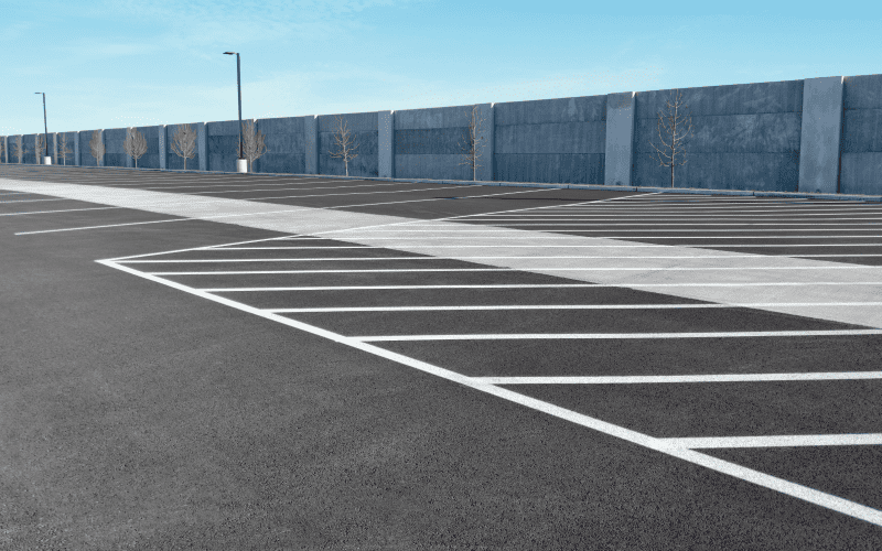 Empty parking lot with white painted lines and a concrete wall in the background under a clear blue sky.