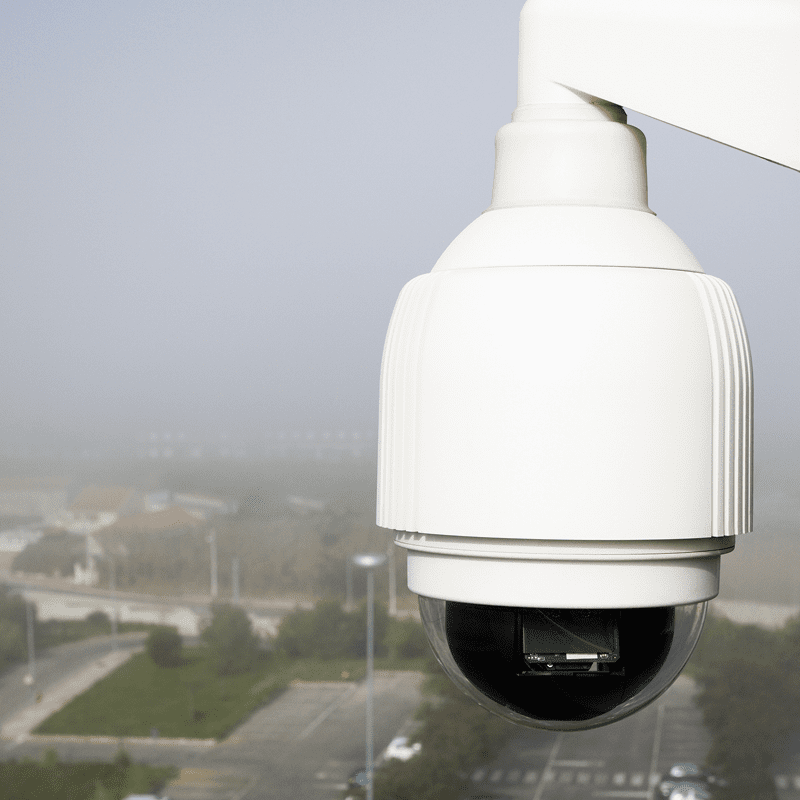 Close-up of a white security camera overlooking a parking lot. The area below is visible but slightly obscured by fog.