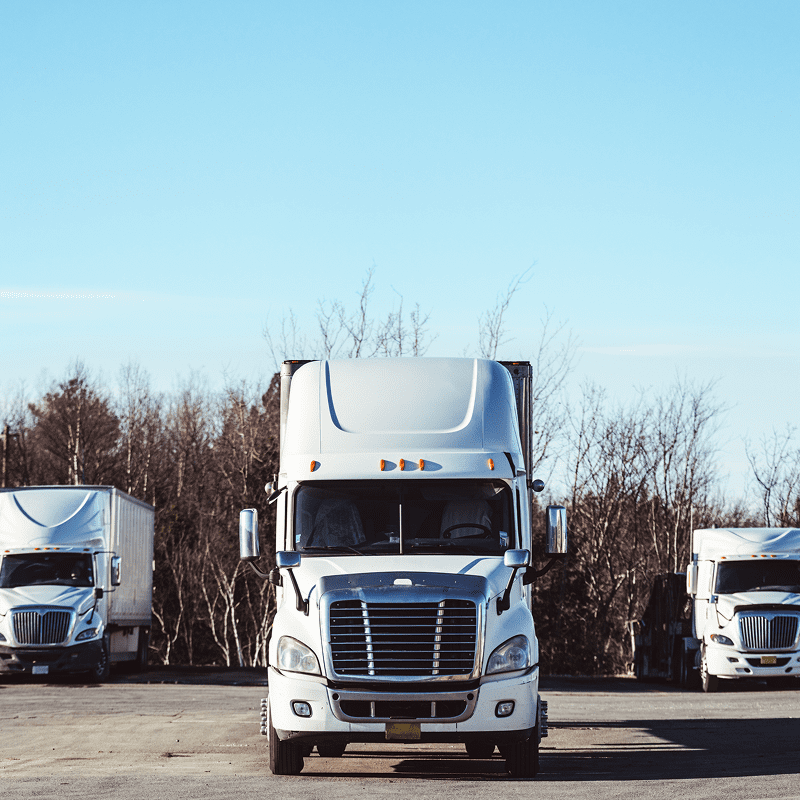 Three white semi-trucks are parked on a paved lot with trees in the background under a clear blue sky.